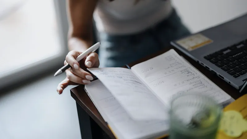 Hands writing in a notebook beside a laptop on a desk
