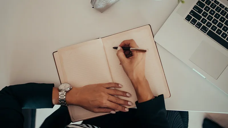 Hands writing notes in a notebook next to an open laptop